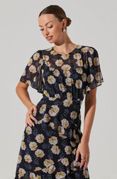 A woman is wearing a black dress with a floral pattern featuring large white, yellow, and lavender flowers with green and purple accents. The dress has short, semi-sheer, ruffled sleeves and a rounded neckline. The fabric appears lightweight and flowy, with a fitted waist.